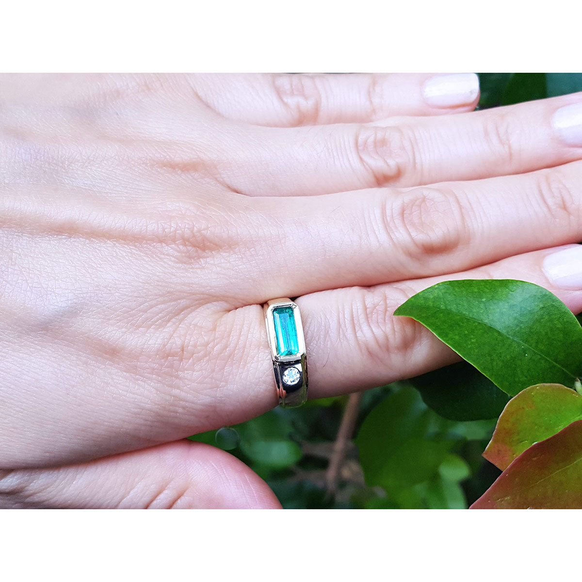 A person&#39;s hand wearing a mens emerald ring in yellow gold on a leafy background.