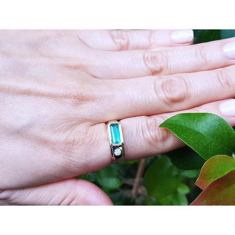 A person's hand wearing a mens emerald ring in yellow gold on a leafy background.