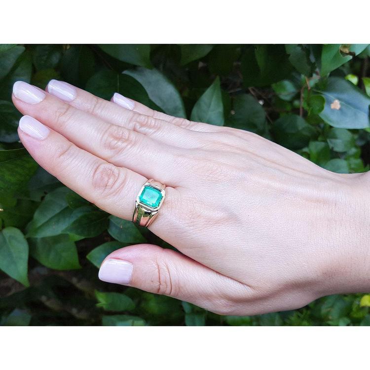 Hand wearing a gold ring for men with a natural emerald against a green leafy background.