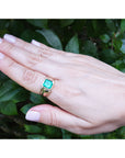 Hand wearing a gold ring for men with a natural emerald against a green leafy background.