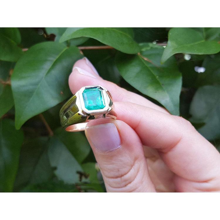 Gold men's emerald ring held by a hand against a leafy background.