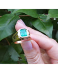 Gold men's emerald ring held by a hand against a leafy background.