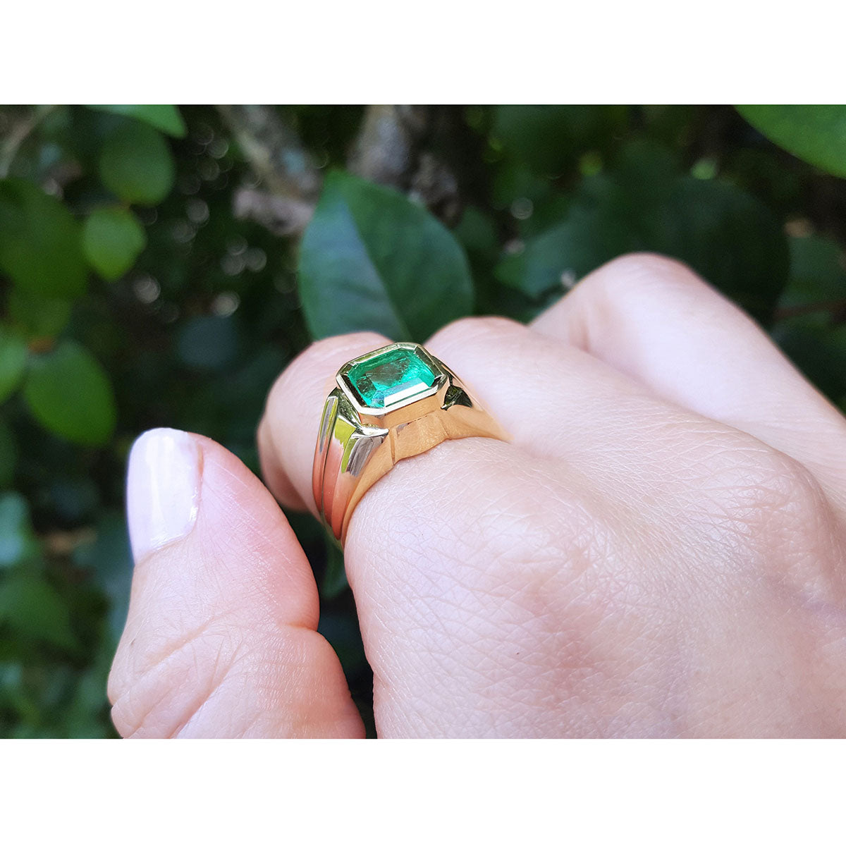 Gold men&#39;s ring with a natural emerald on a hand against a blurred green leafy background.