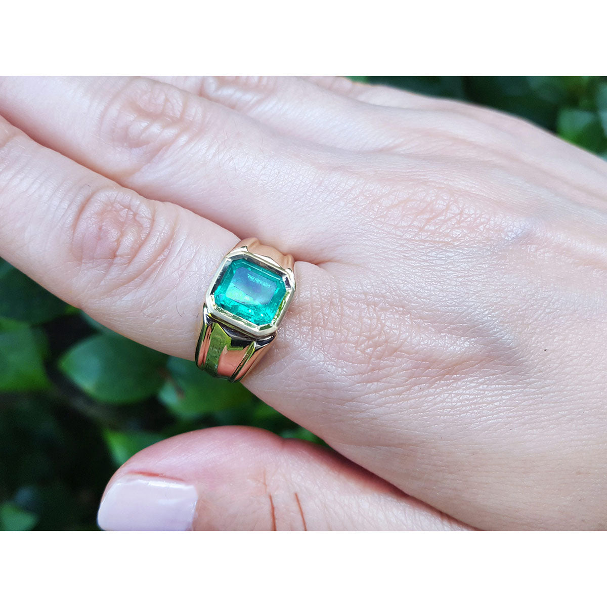 Gold ring for man with a square natural emerald on a hand against a blurred green background.