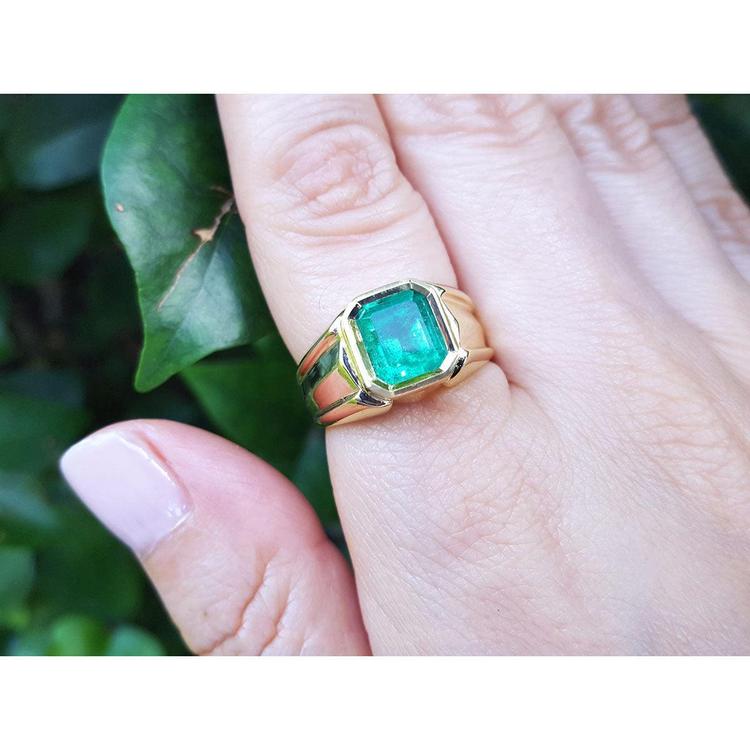 A yellow gold solitaire ring for men with a square emerald bezel set on a hand against a natural background.
