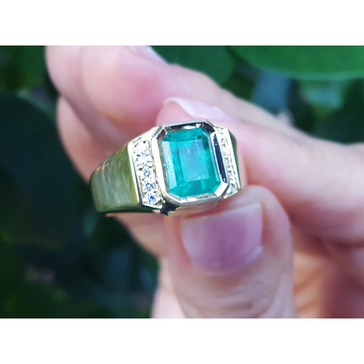 Gold ring with a natural emerald held between fingers against a blurred natural background.