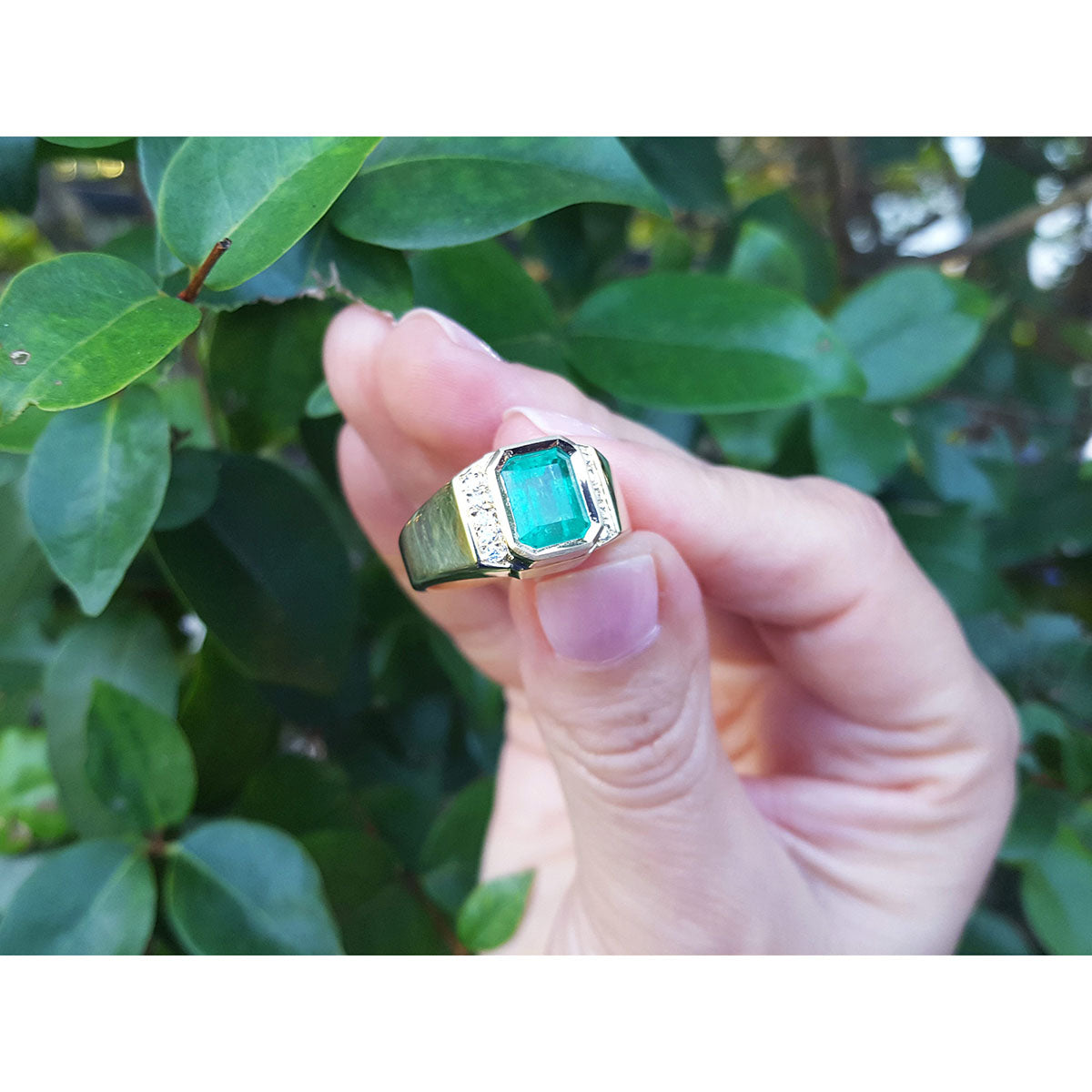 Hand holding a ring with natural emerald and diamonds accent against a green leafy background.