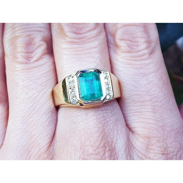 
This 18k yellow gold men's emerald ring is in bezel set and has accent natural diamonds on a person's finger.