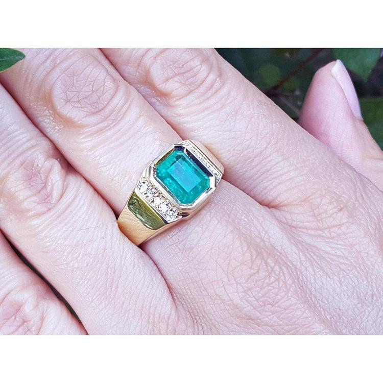 An emeral-cut men's emerald ring in yellow gold with natural accent diamonds on a person's finger.