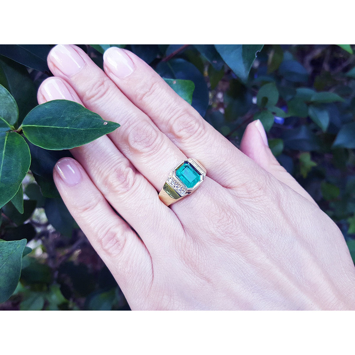 A close view of a hand wearing a natural colombian emerald ring for men against a leafy background.