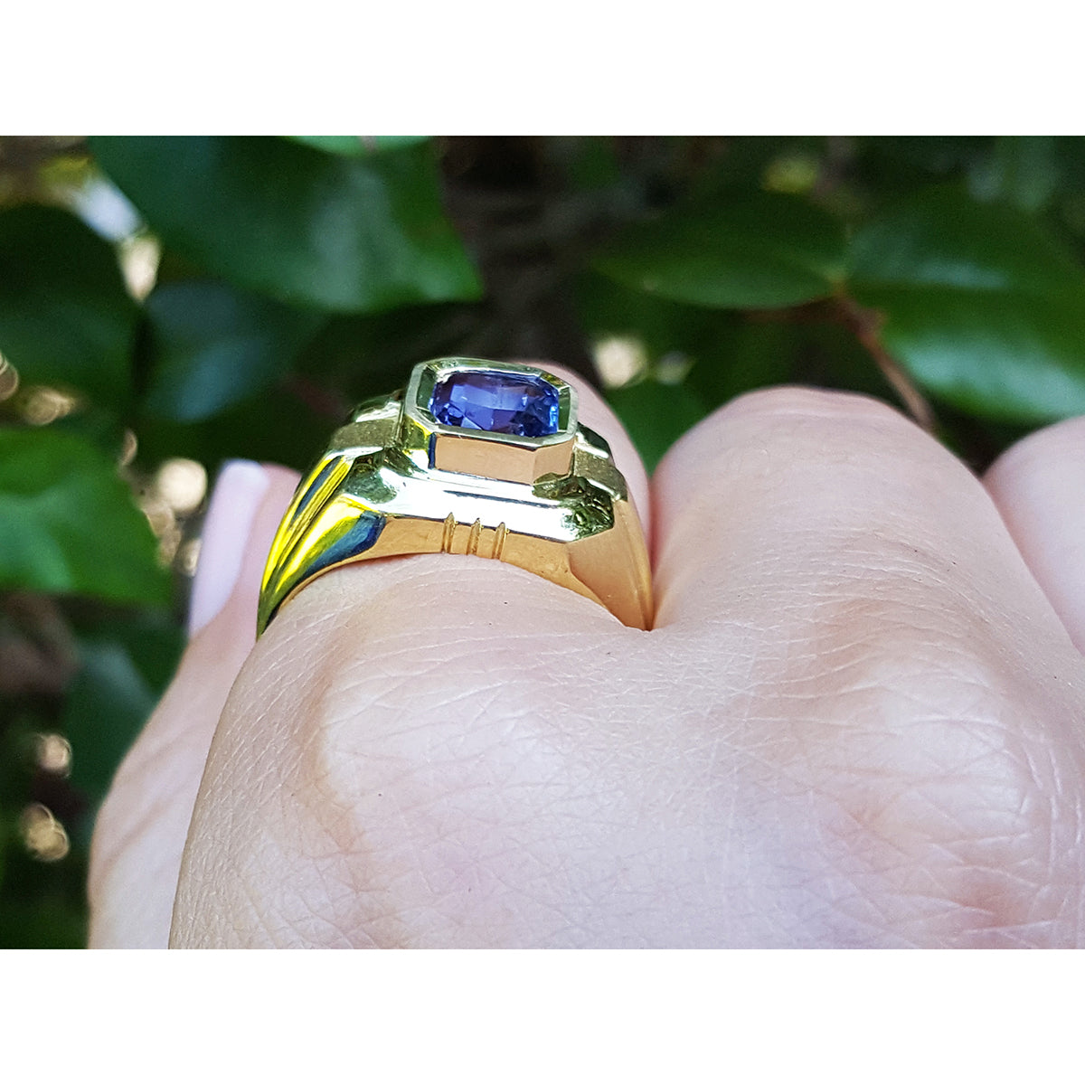 Gold ring with a blue natural sapphire sagemstone on a finger against a blurred green leafy background