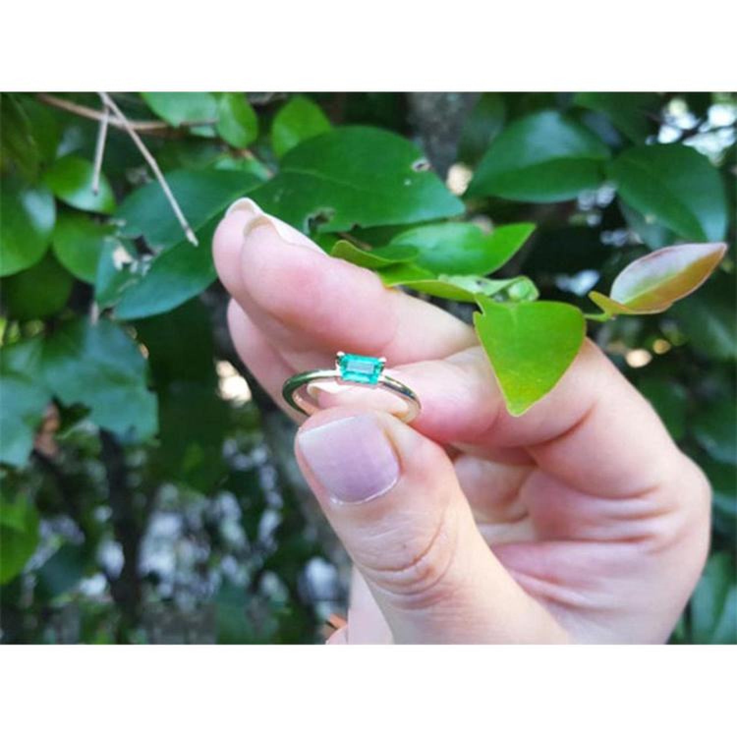Minimalist 0.35 ct. emerald solitaire ring, east-west set in 14K yellow gold, held against green leaves.