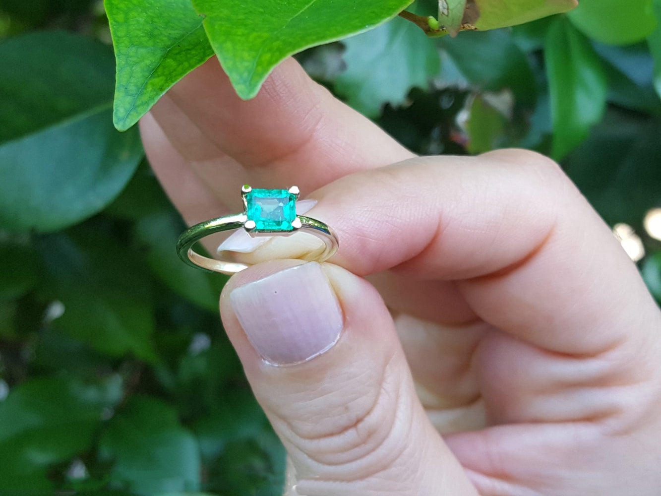 A minimalist emerald solitaire ring held between fingers against a green leafy background.