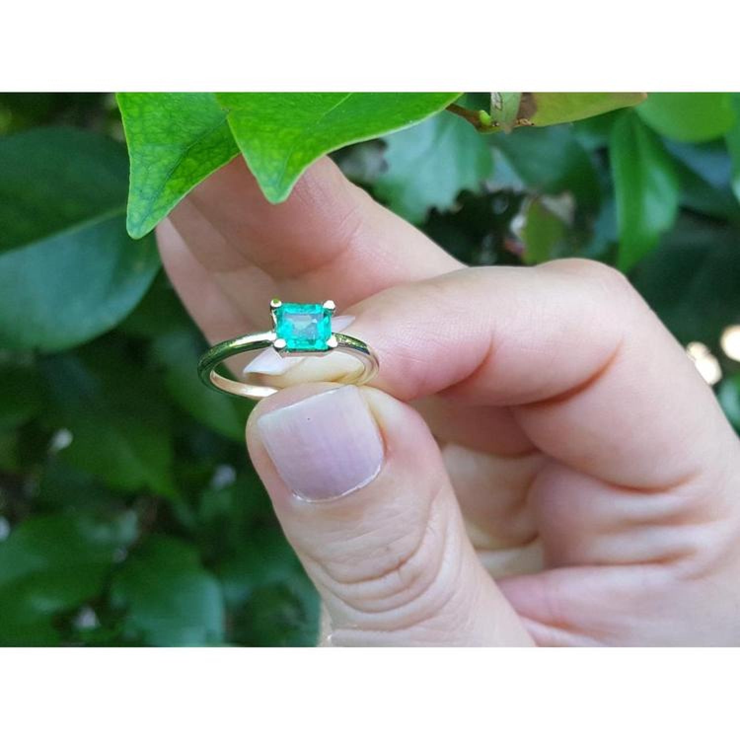 Minimalist 14K yellow gold ring with a square-cut natural Colombian emerald, held by fingers near green leaves.