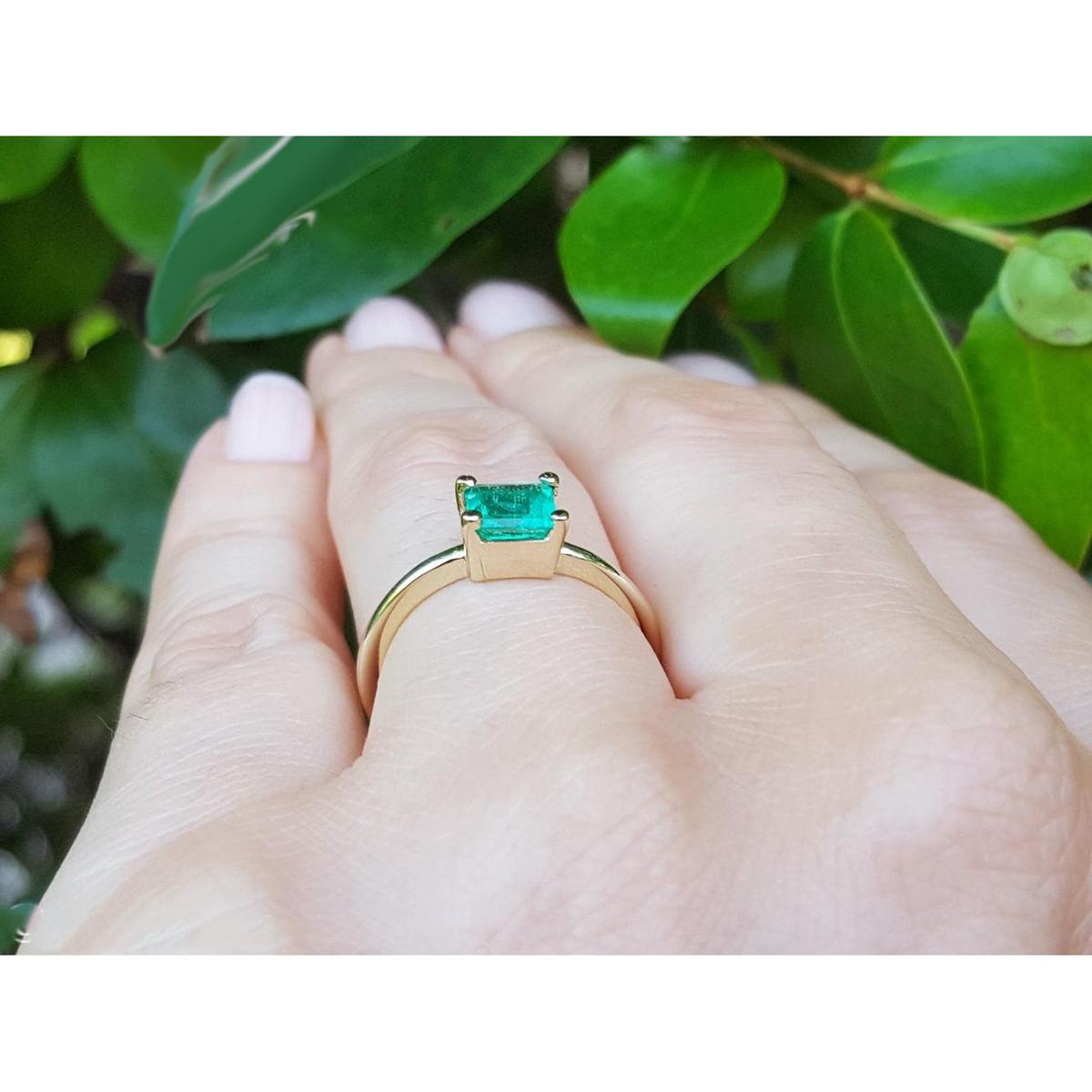A solitaire emerald gold ring on a hand against a leafy background.