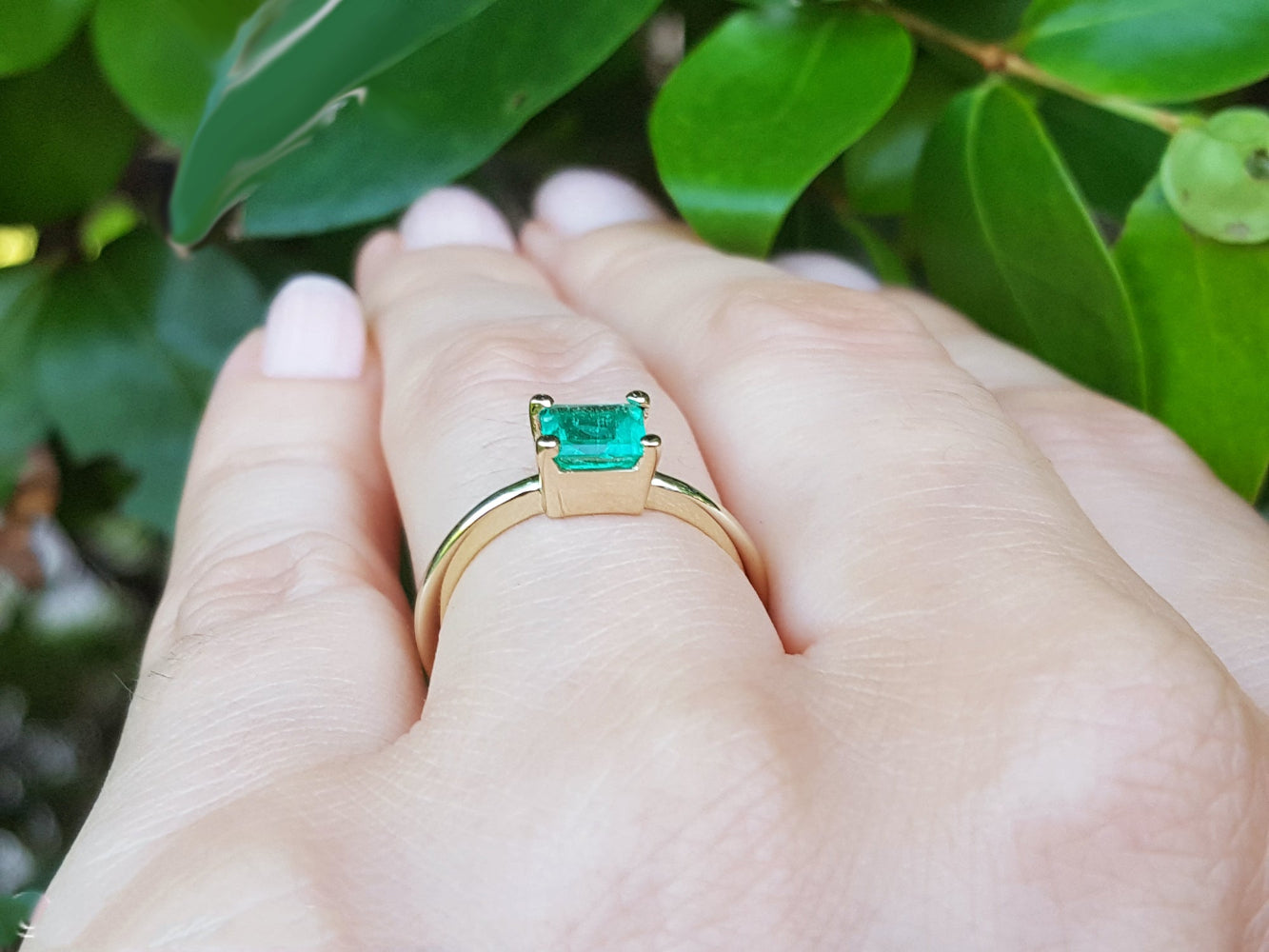 A solitaire emerald gold ring on a hand against a leafy background.