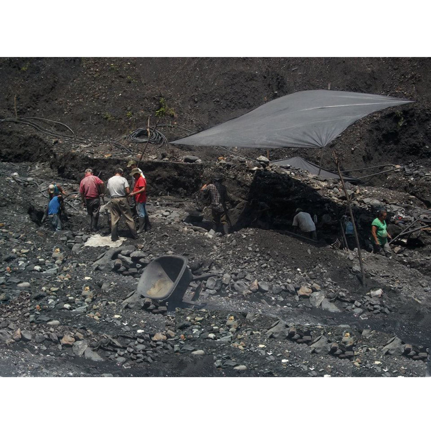 Searching for emeralds. A group of miners' people standing and working around a dark opening or tunnel in a rocky, debris-filled outdoor site under a large tarp canopy.