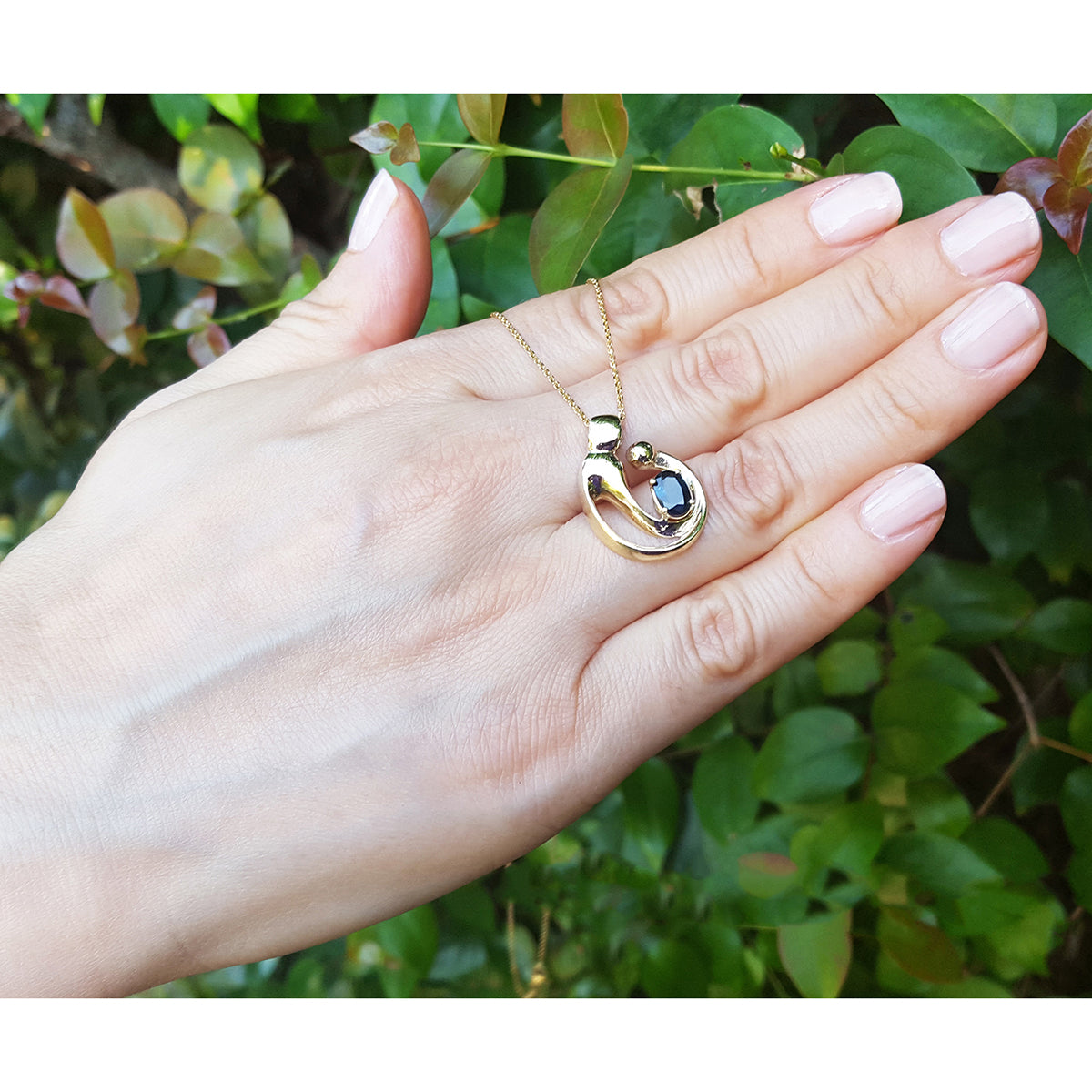 Hand displaying a yellow gold necklace with a mother and child pendant against a green leafy background