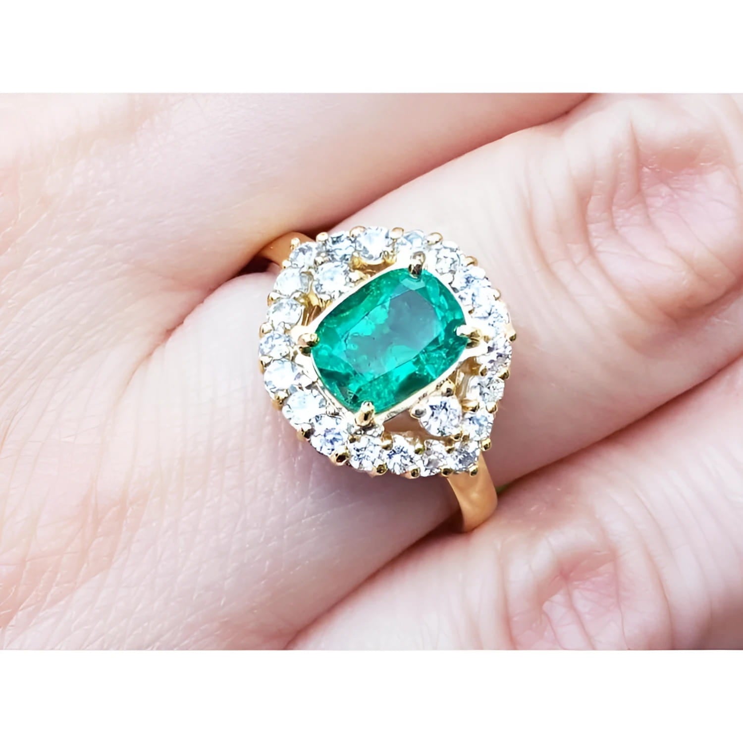 A cushion cut Muzo colombian emerald surrounded by a halo diamond 14k gold ring on a person's finger.