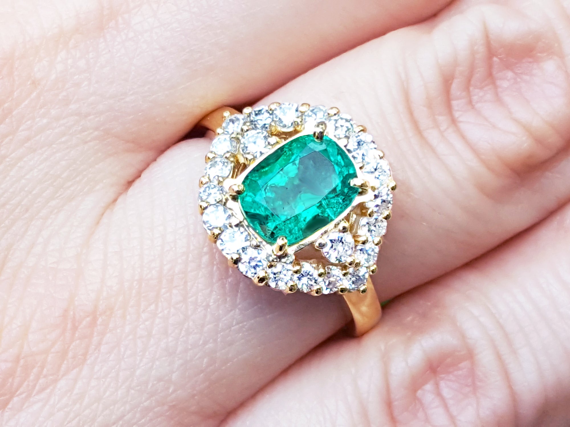 A cushion cut Muzo colombian emerald surrounded by a halo diamonds ring on a person's finger.