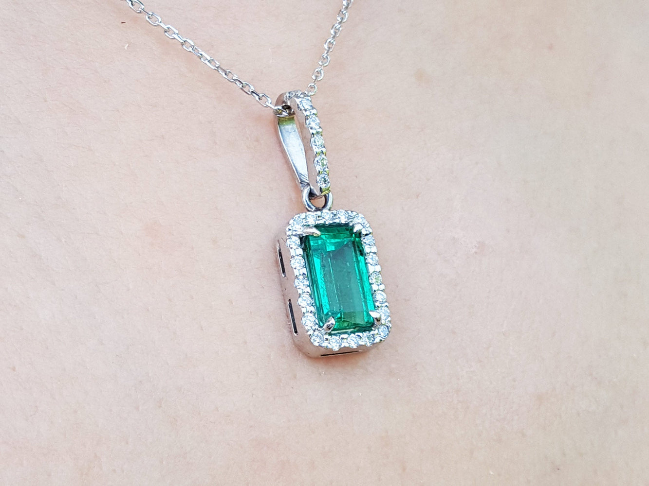 A natural emerald pendant on a silver white gold chain against a beige skin background