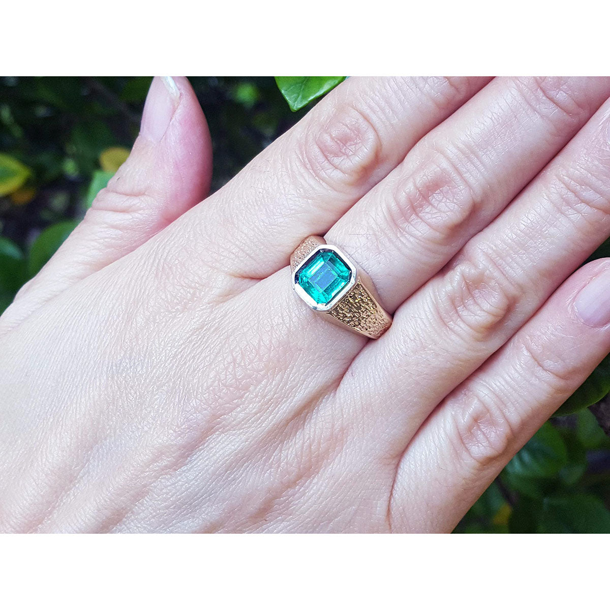 Hand wearing a ring with a square emerald against a blurred natural background.