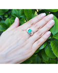 Hand wearing a men's solitaire ring with a Muzo colombian emerald against a leafy background.
