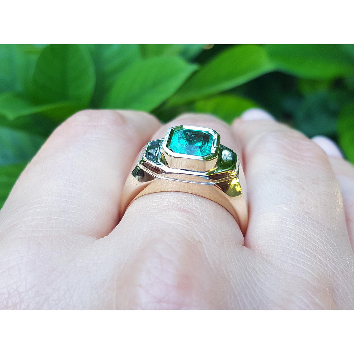 Gold ring with a genuine emerald on a hand against a green leafy background.