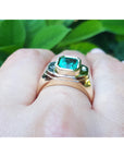 Gold ring with a genuine emerald on a hand against a green leafy background.
