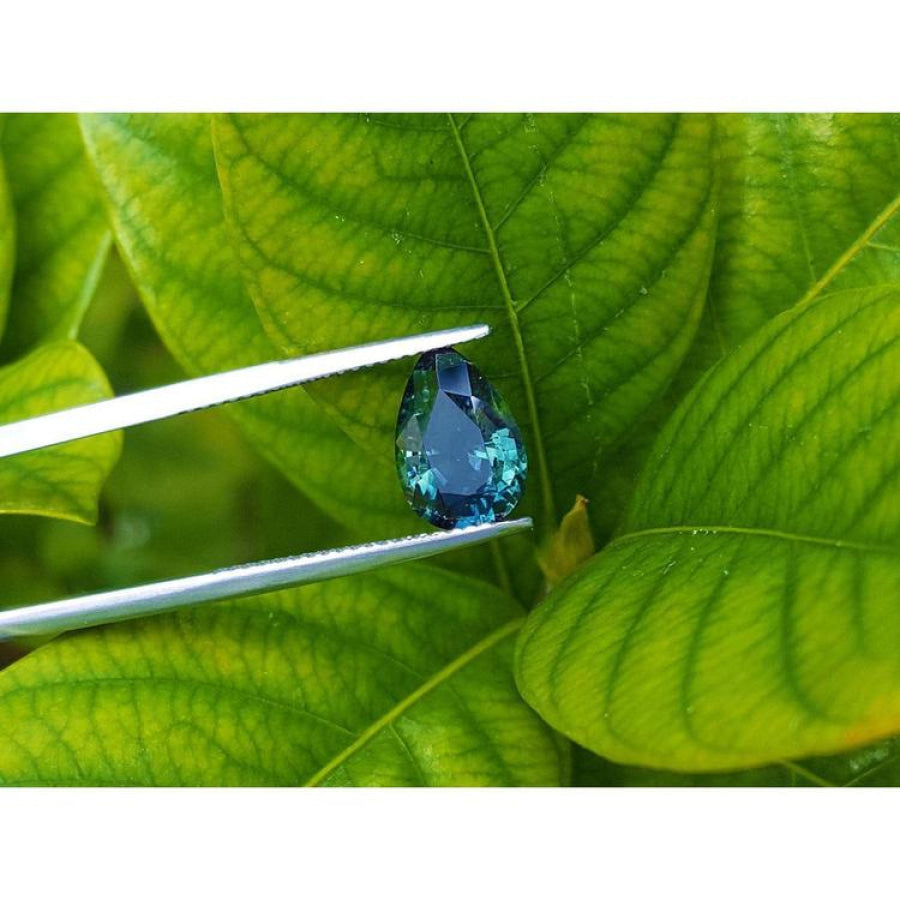 3.22 ct. loose Australian blue sapphire, pear-shaped, deep blue-green, held by tweezers against green leaves.