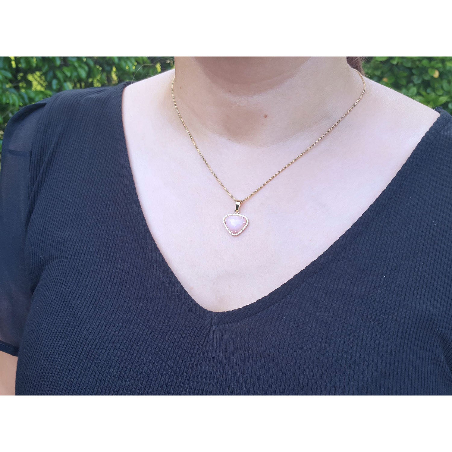 A woman wearing a gold necklace with a heart-shaped opal pendant on a blue top against a green background.