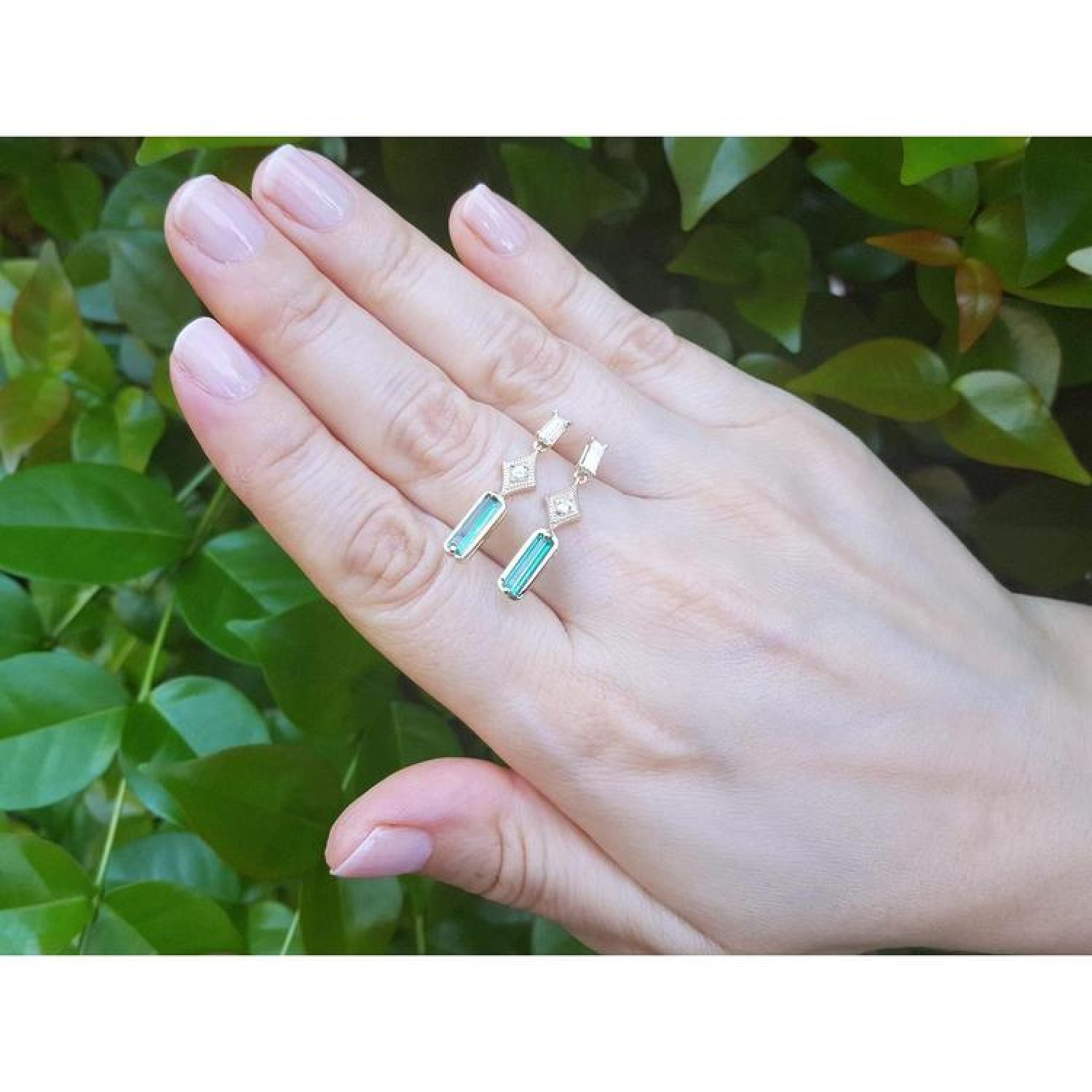 1.05 carat natural baguette cut emerald earrings in 14K gold, held against green foliage.