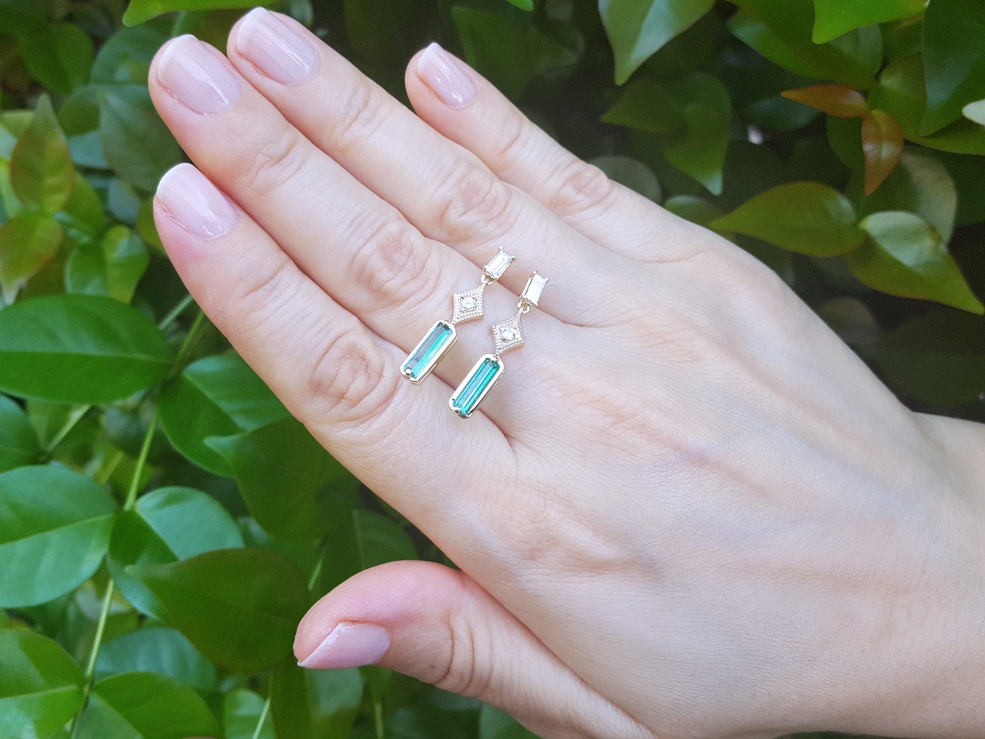 Hand wearing a pair of emerald earrings with geometric design against a green leafy background