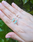 Hand wearing a pair of emerald earrings with geometric design against a green leafy background