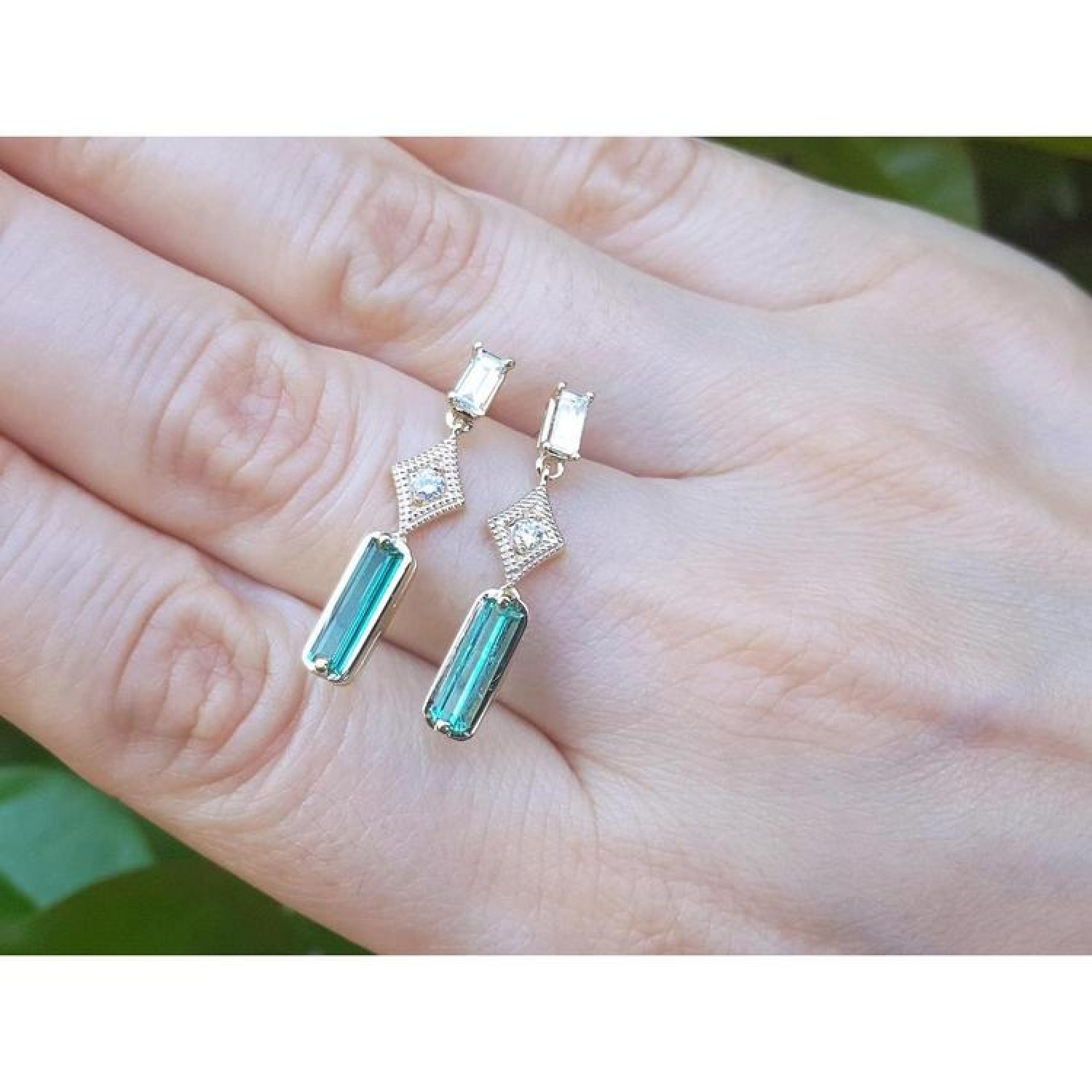 14K gold baguette emerald drop earrings with diamond accents on a hand.