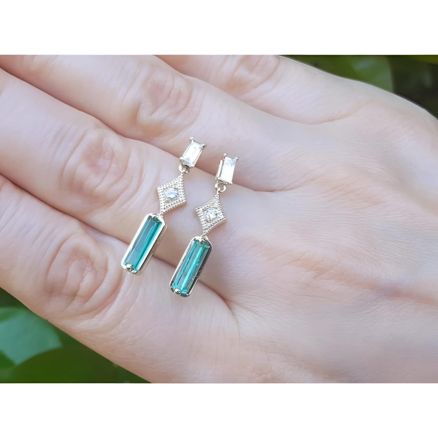 14K gold baguette emerald drop earrings with diamond accents on a hand.