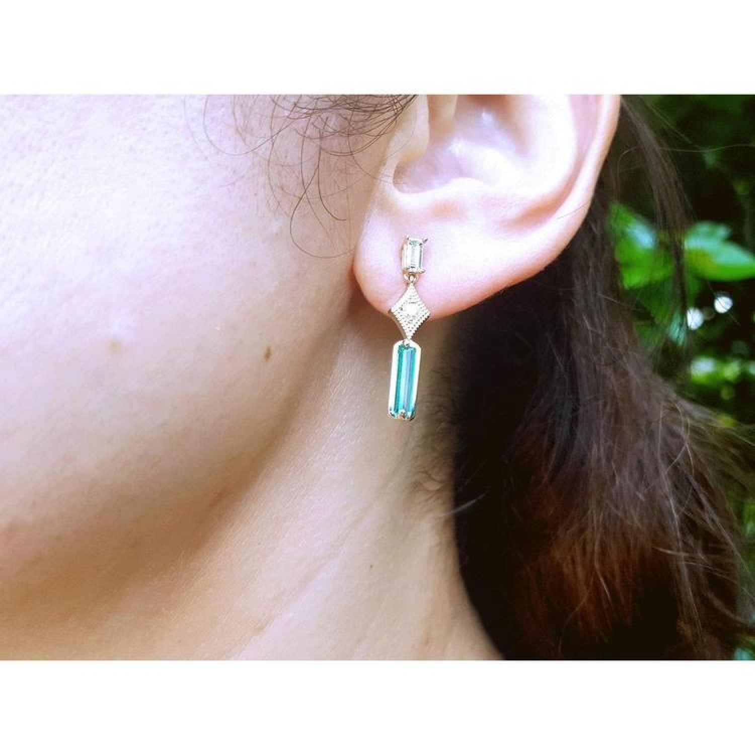 14K gold baguette cut emerald drop earrings with diamond accents on a woman's ear.