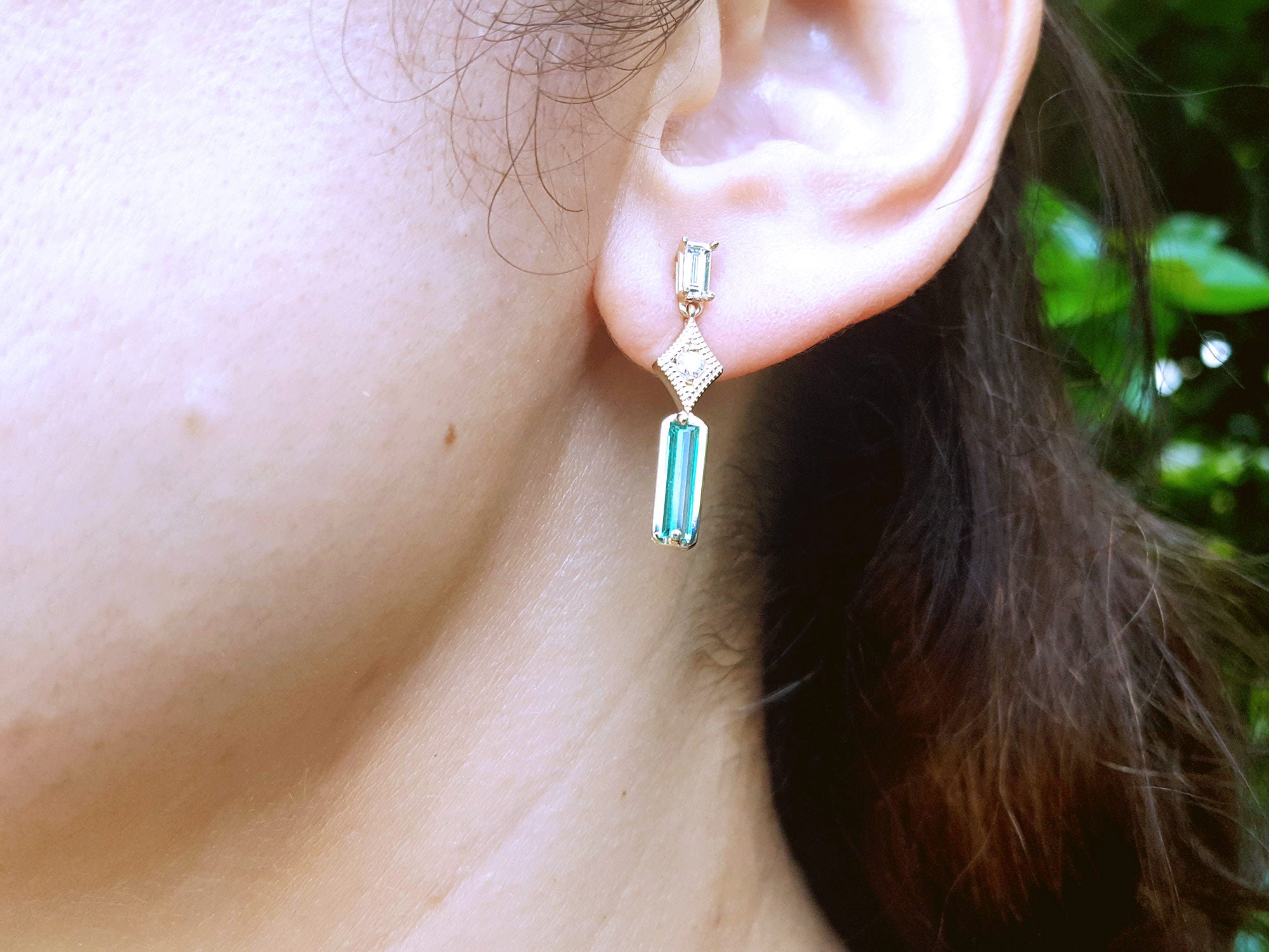 Close-up of an ear wearing a gold emerald and diamond earring with a blurred green background