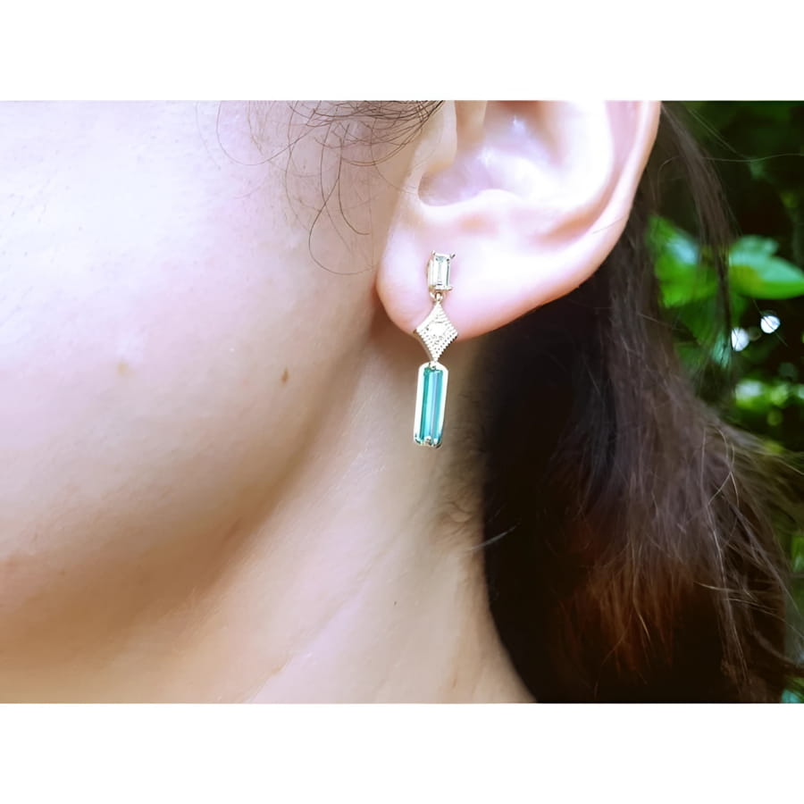 14K gold baguette cut emerald drop earrings with diamond accents on a woman's ear.