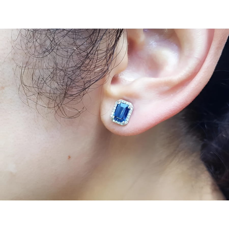 Emerald cut natural blue sapphire stud earring with diamond halo in 14K white gold on an ear.