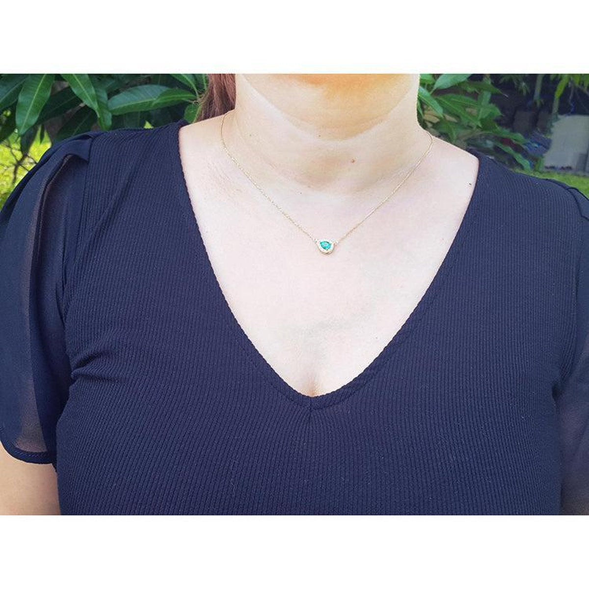 Woman wearing a vibrant pear-shaped emerald necklace with diamond halo on a 14K yellow gold chain.