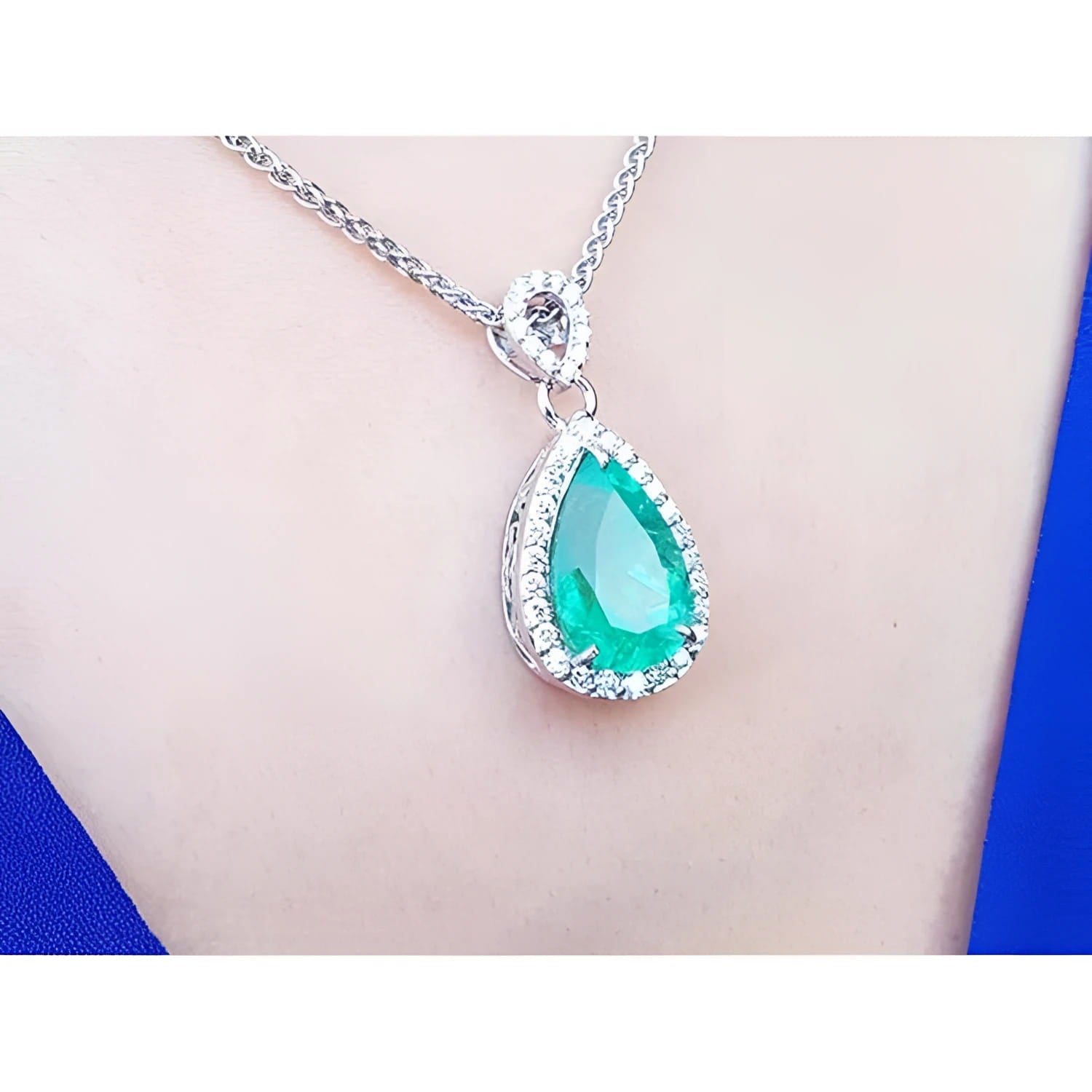 Pear-cut Colombian emerald pendant with diamond halo on 18K white gold chain, worn on a blue top.