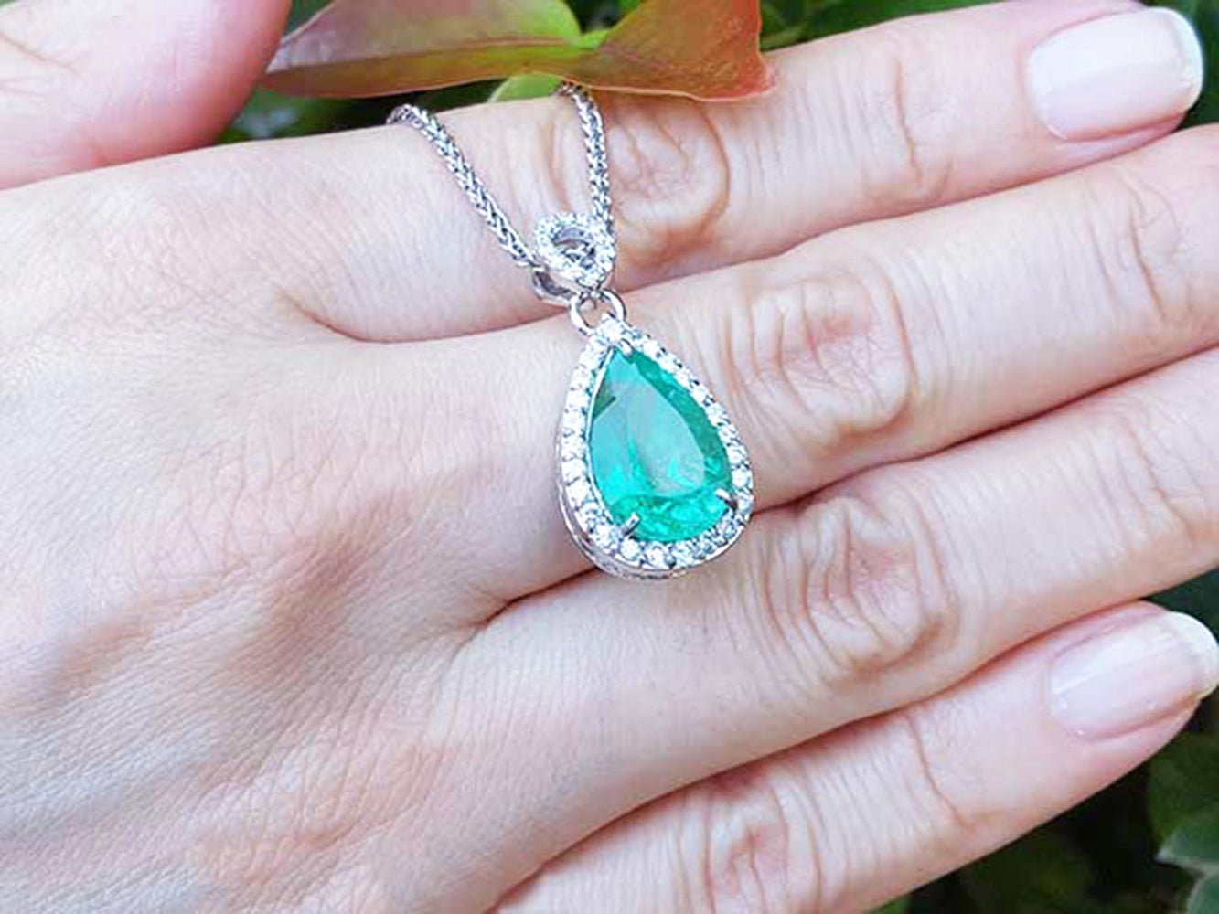Green teardrop emerald pendant on a gold chain held on a hand with a blurred green background.