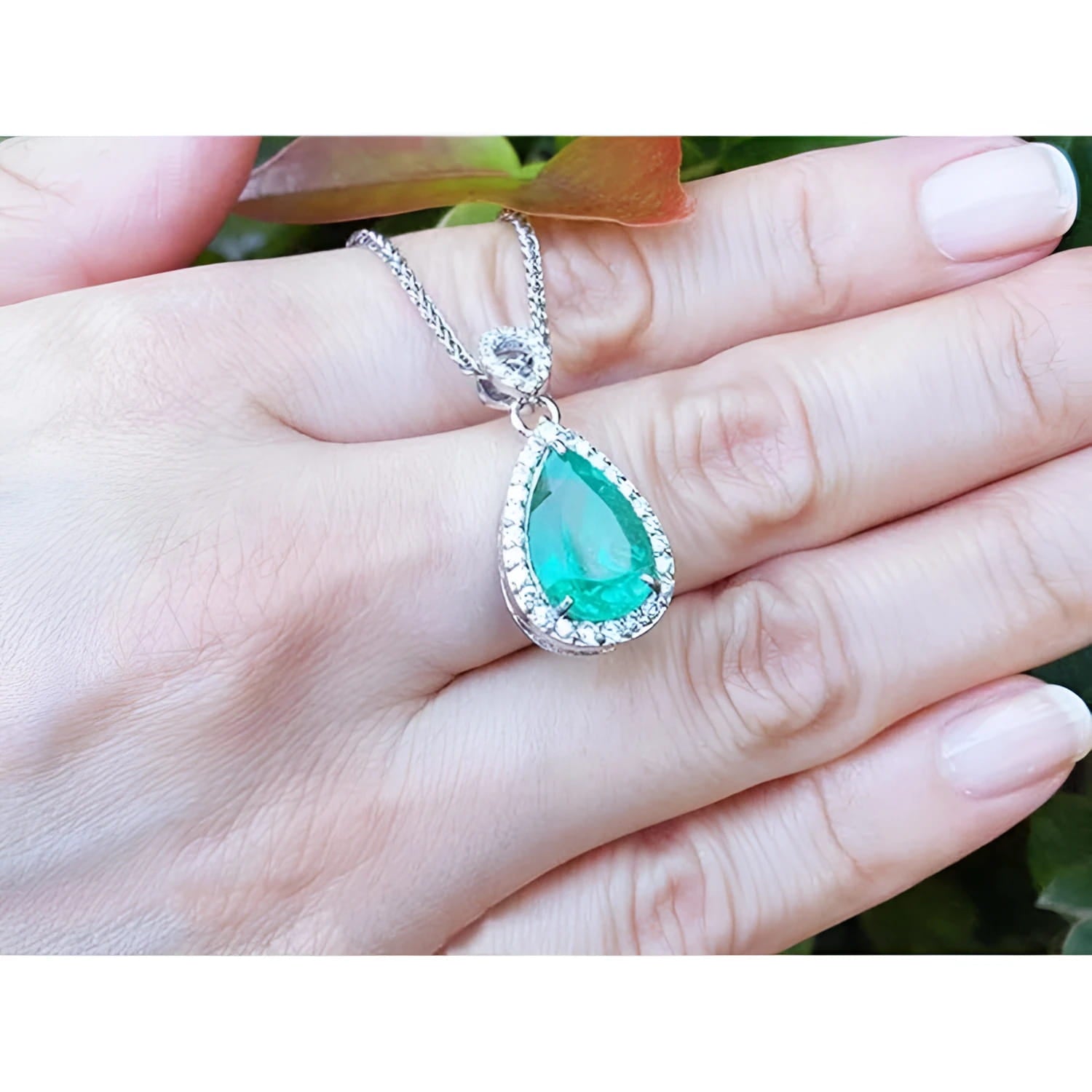 5.83 ct. Colombian emerald pear cut pendant with diamond halo on 18K white gold chain, held in hand.