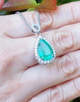 Green teardrop emerald pendant on a gold chain held on a hand with a blurred green background.