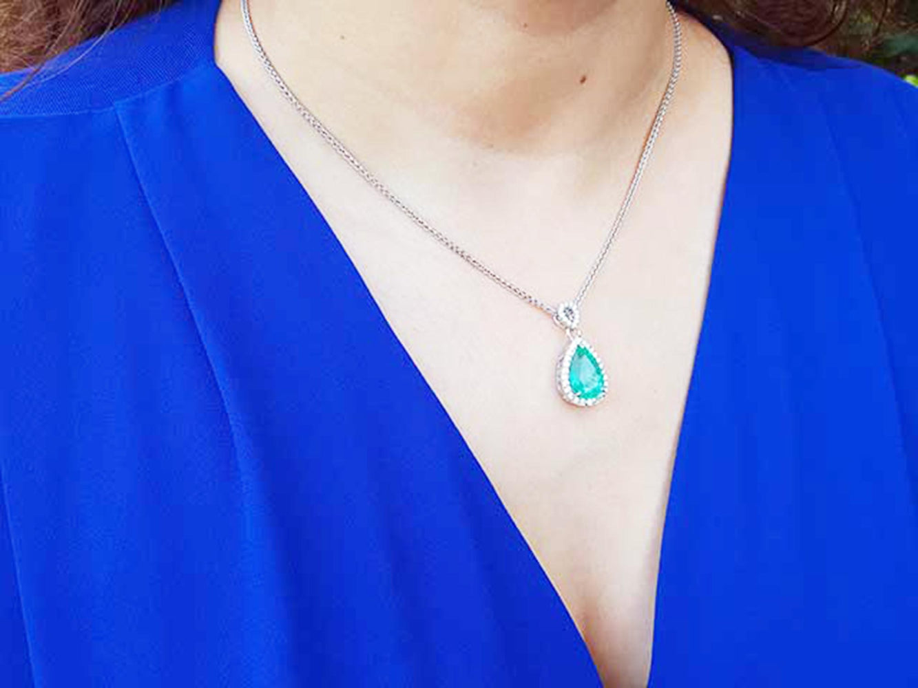 A woman wearing a blue top with a necklace featuring a teardrop emerald pendant.