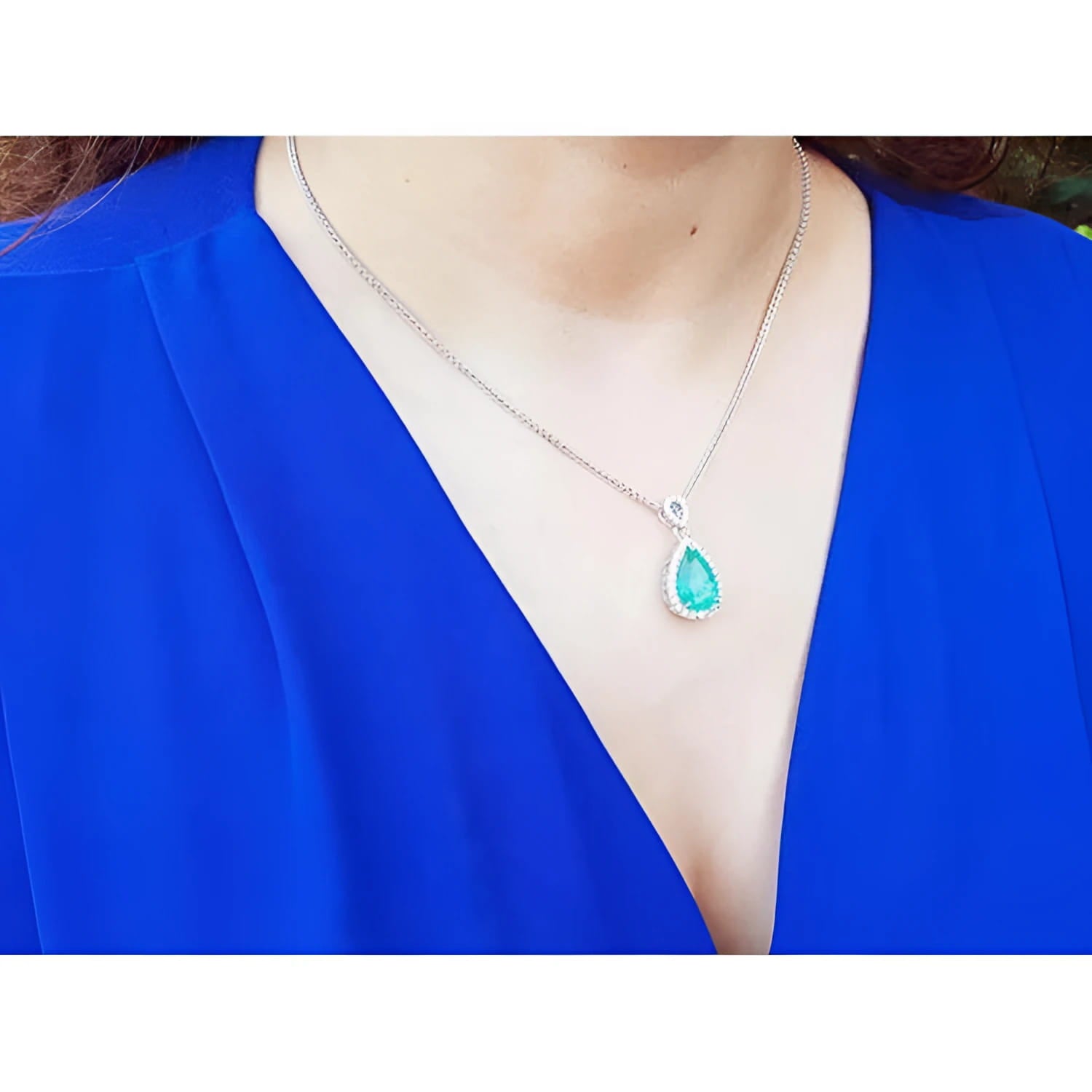 Pear-cut Colombian emerald pendant with diamond halo on an 18K white gold chain, worn on a blue top.