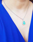A woman wearing a blue top with a necklace featuring a teardrop emerald pendant.