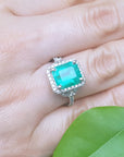 A green fire emerald ring on a hand with green leaves in the background.
