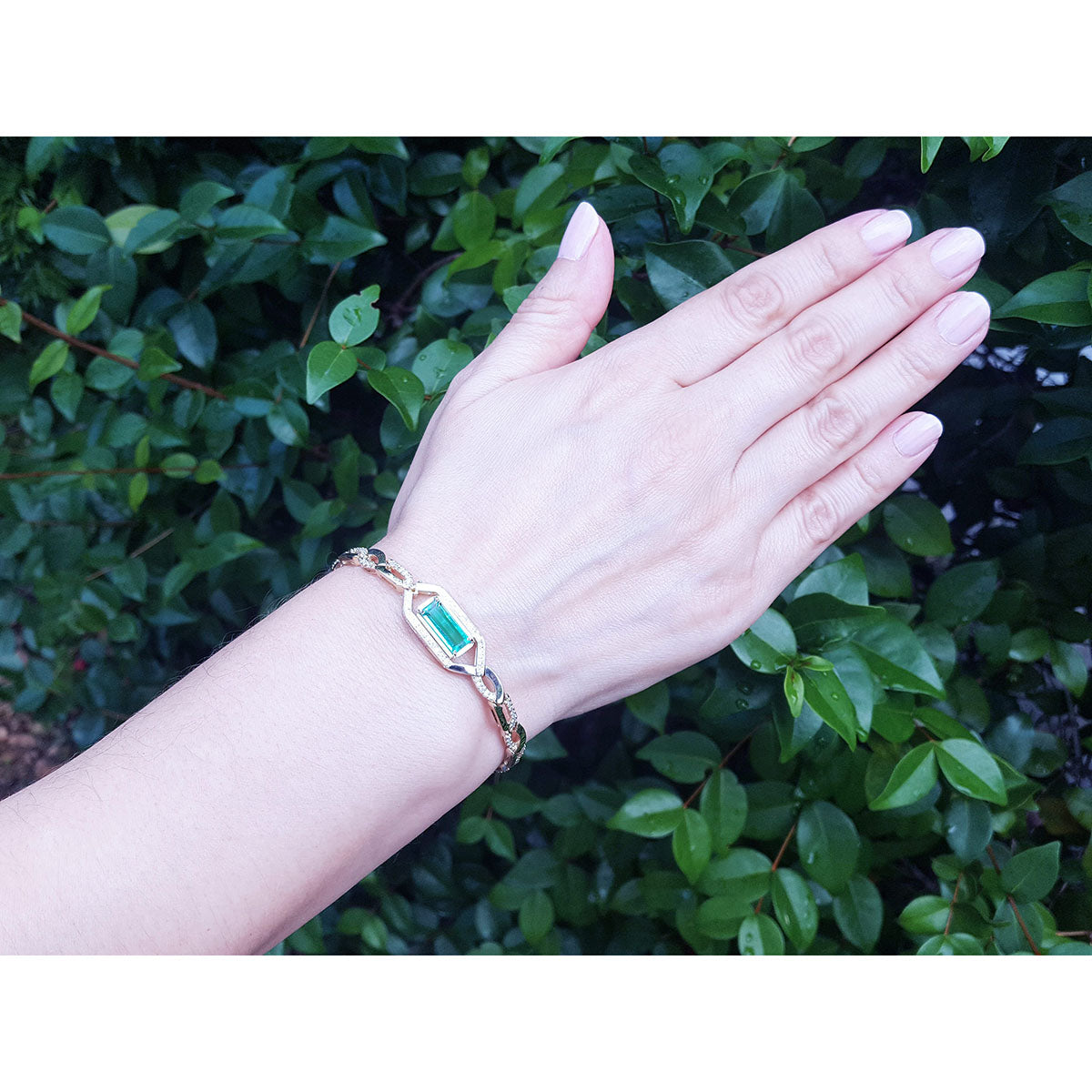 Hand wearing a bracelet with a natural emerald gemstone against a green leafy background